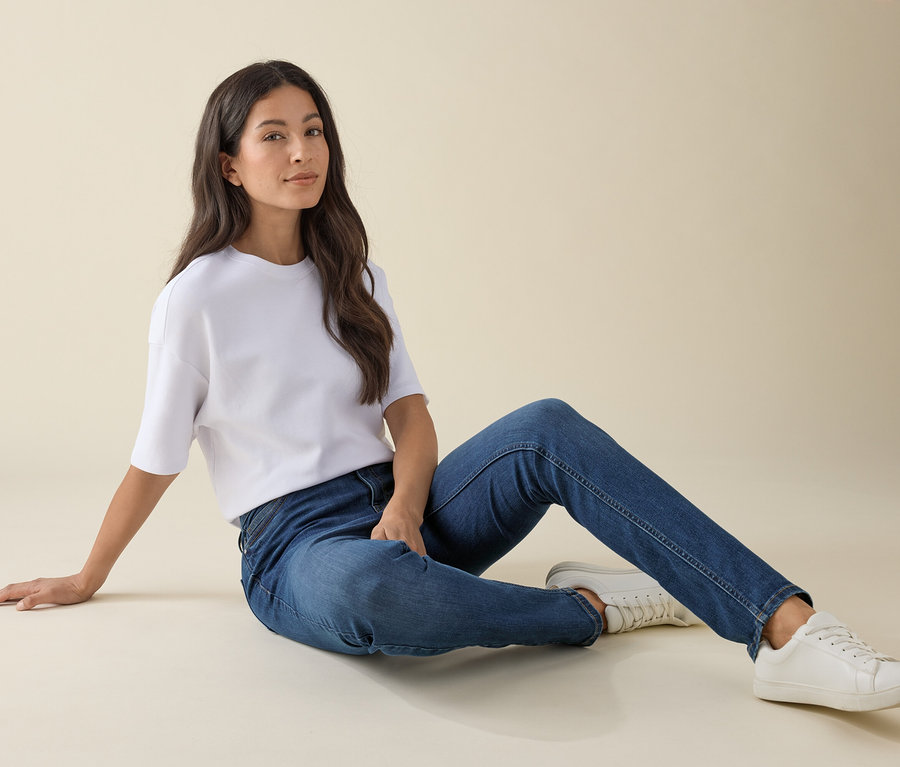 Žena sedí na zemi v slim džínsoch – strih »Emma«, modrý denim, bielom tričku a teniskách.