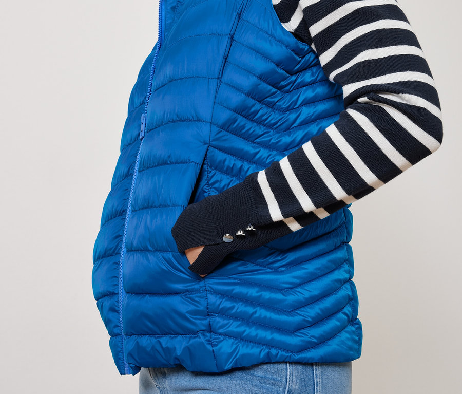 Osoba má ruku vo vrecku modrej prešívanej vesty a má na sebe námornícke modro-biele pruhované tričko.