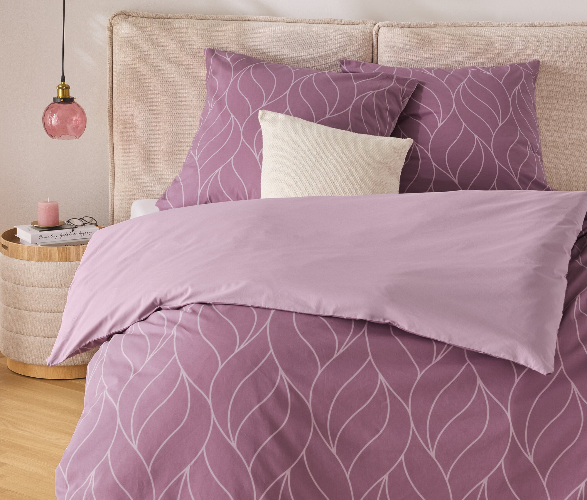 Fialové posteľné obliečky s bielym vzorom listov na posteli s béžovým čelom. Nočný stolík s lampou stojí vedľa postele.