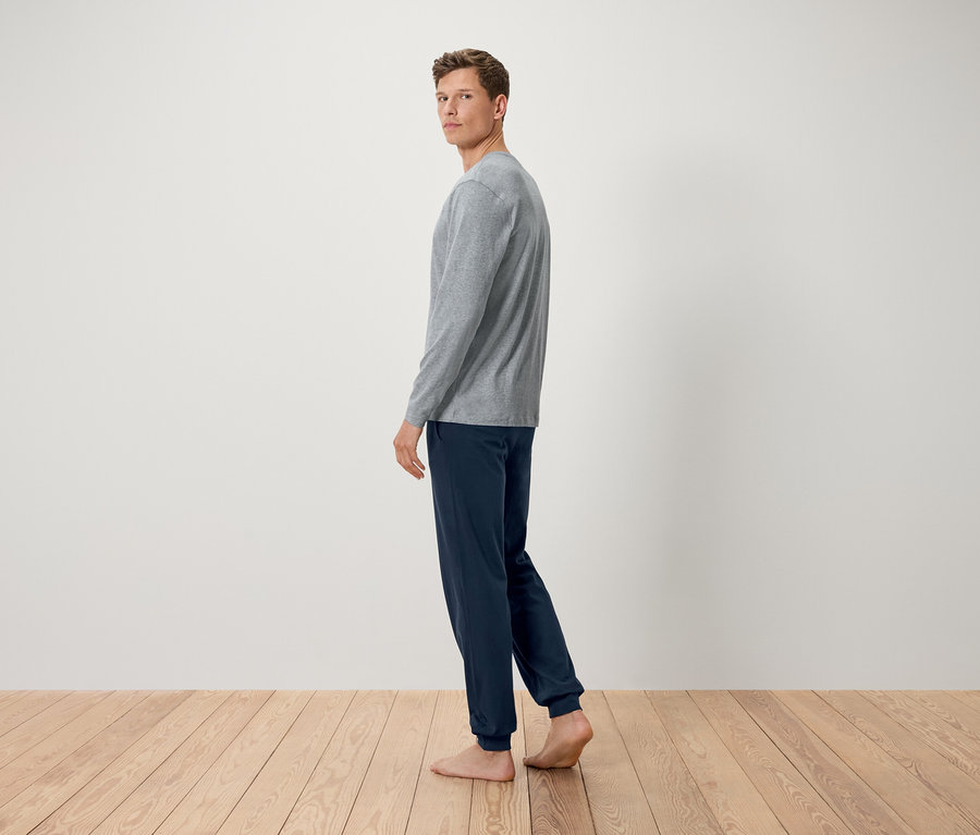 Muž v sivom tričku s dlhým rukávom a tmavomodrých nohaviciach stojí bosý na drevenej podlahe a pozerá sa cez plece.