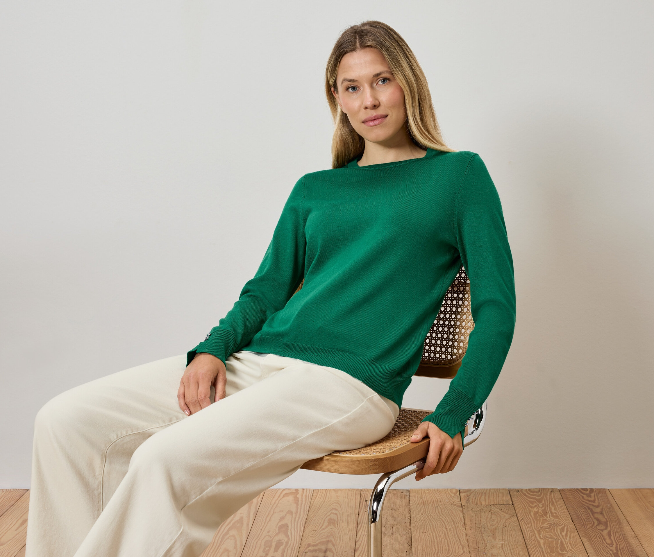 Blondínka sedí na stoličke a má na sebe zelený sveter a krémové nohavice.