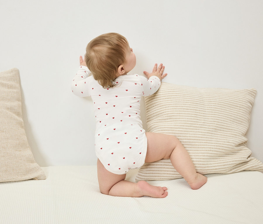 Batoľa v body s dlhým rukávom s červenými srdiečkami stojí pri stene vedľa pruhovaného vankúša.