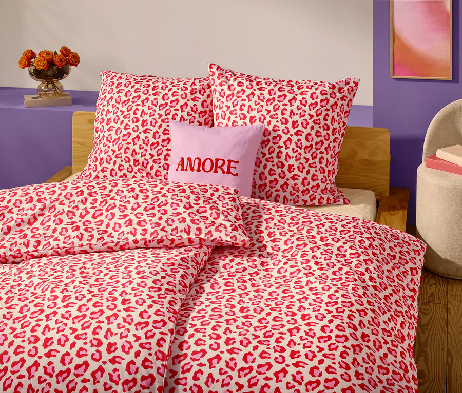 Detailný záber na posteľ s červeno-bielou posteľnou bielizňou s leopardím vzorom a vankúšmi. Levanduľový vankúš s nápisom "AMORE" je umiestnený pred dvoma vankúšmi.