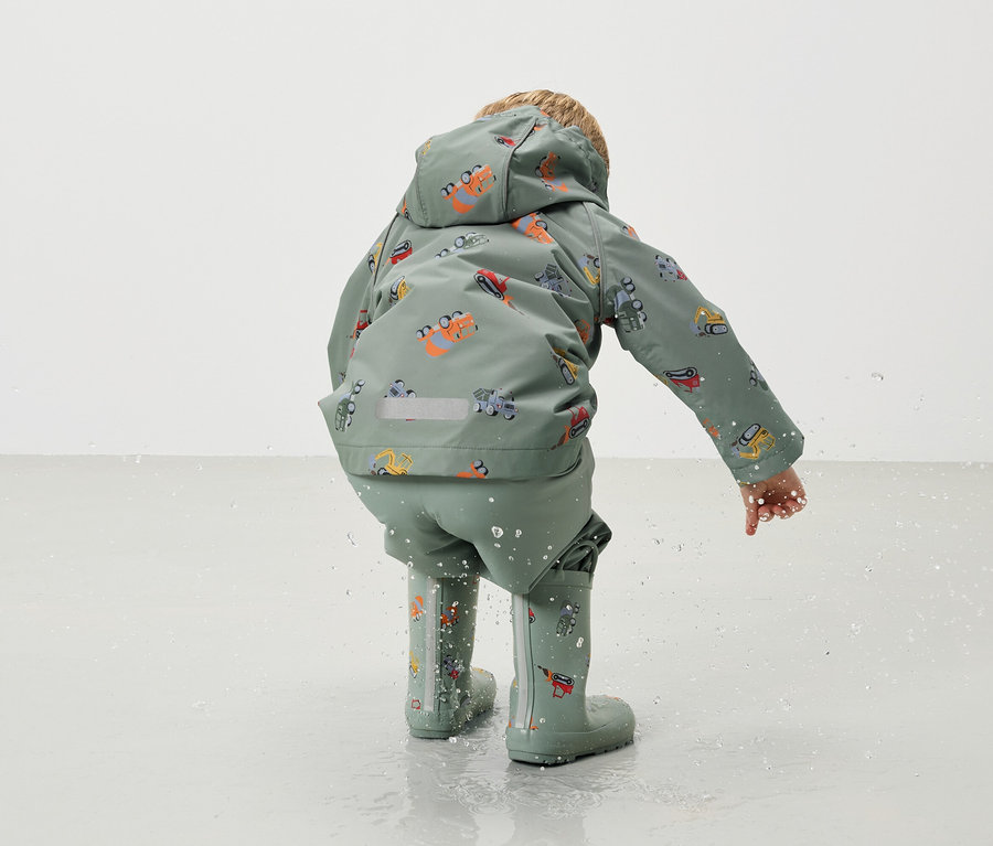 Dieťa zozadu, má na sebe zelenú bundu do dažďa a nohavice do dažďa s motívom bagra a zodpovedajúce gumáky, pričom sa hrá vo vode.