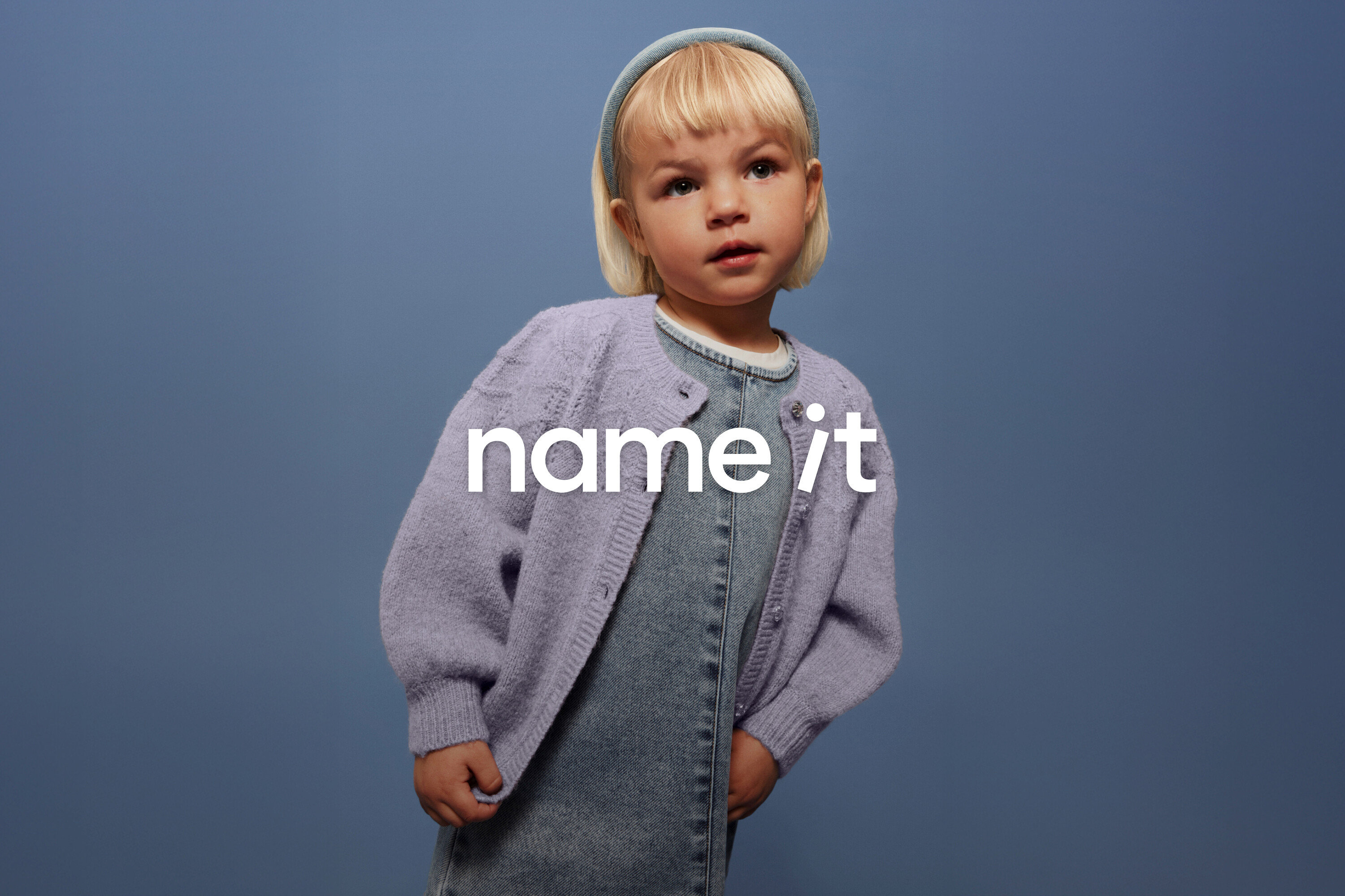 Malé dieťa s blond vlasmi a modrou čelenkou má na sebe džínsové traky a fialový kardigán na modrom pozadí.
