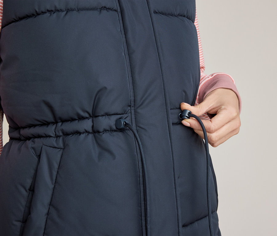 Detail osoby v tmavomodrej prešívanej veste cez ružové pruhované tričko, ktorá si pravou rukou upravuje sťahovaciu šnúrku v páse vesty.