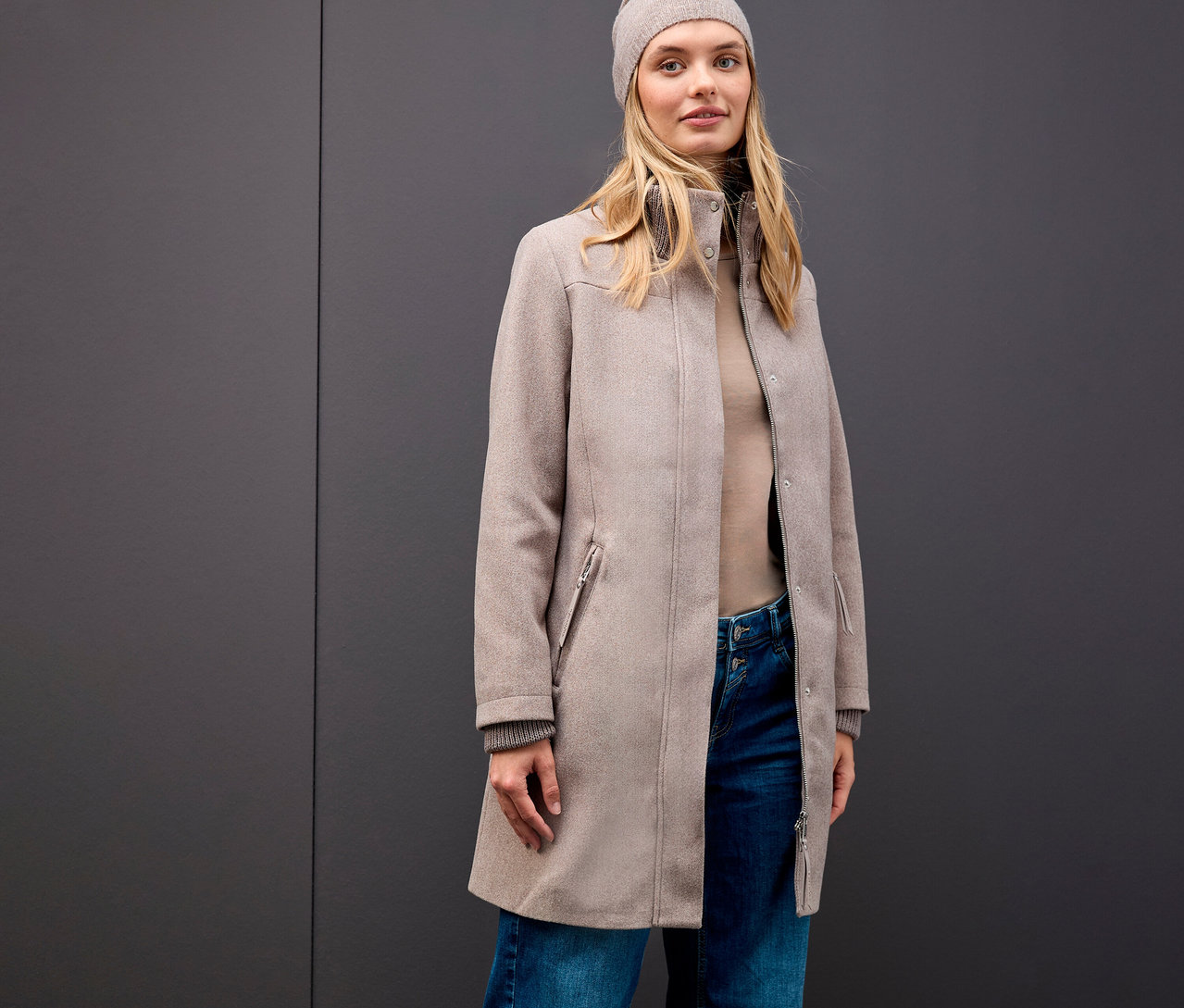 Blondínka má na sebe krátky kabát Street One, čiapku, rolák a džínsy na sivom pozadí.