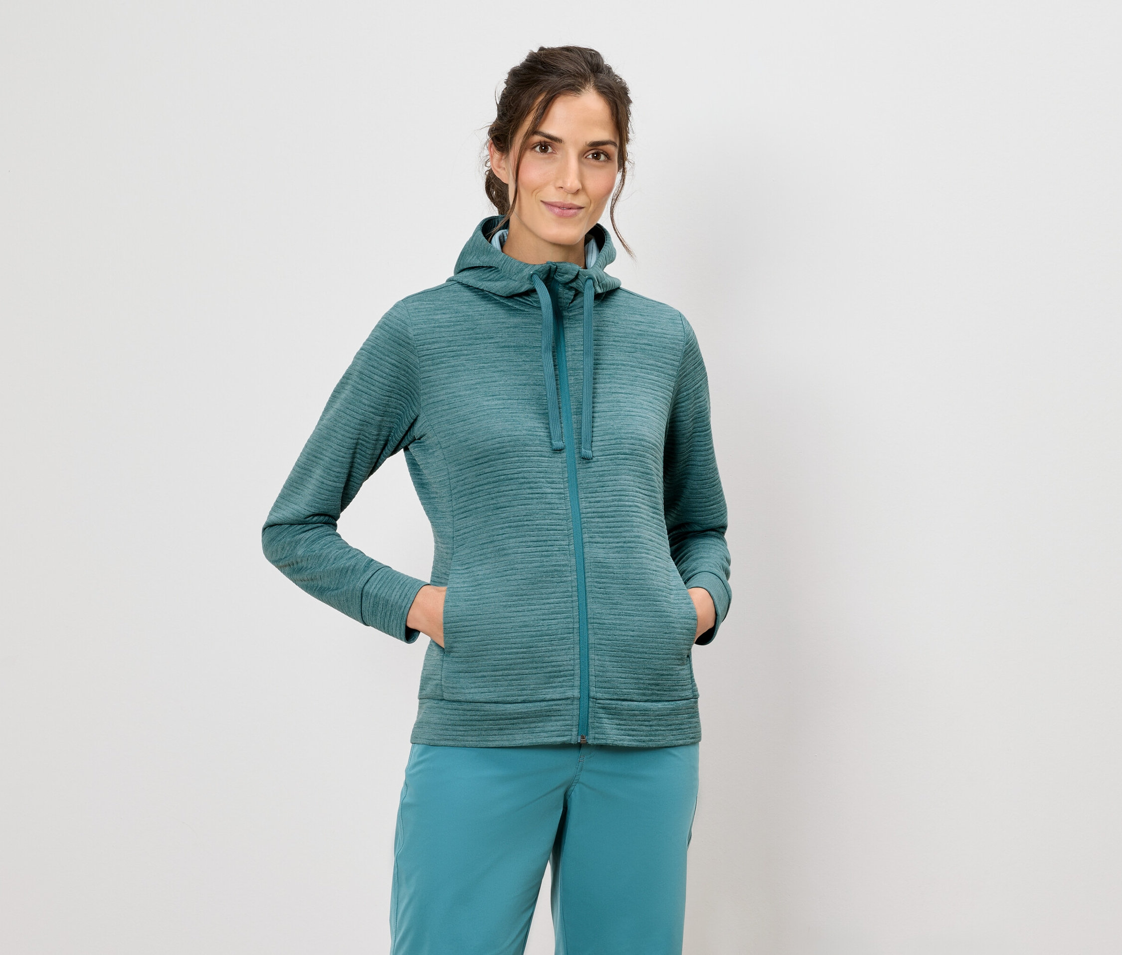 Žena v mikine s kapucňou a modrých nohaviciach stojí na bielom pozadí.