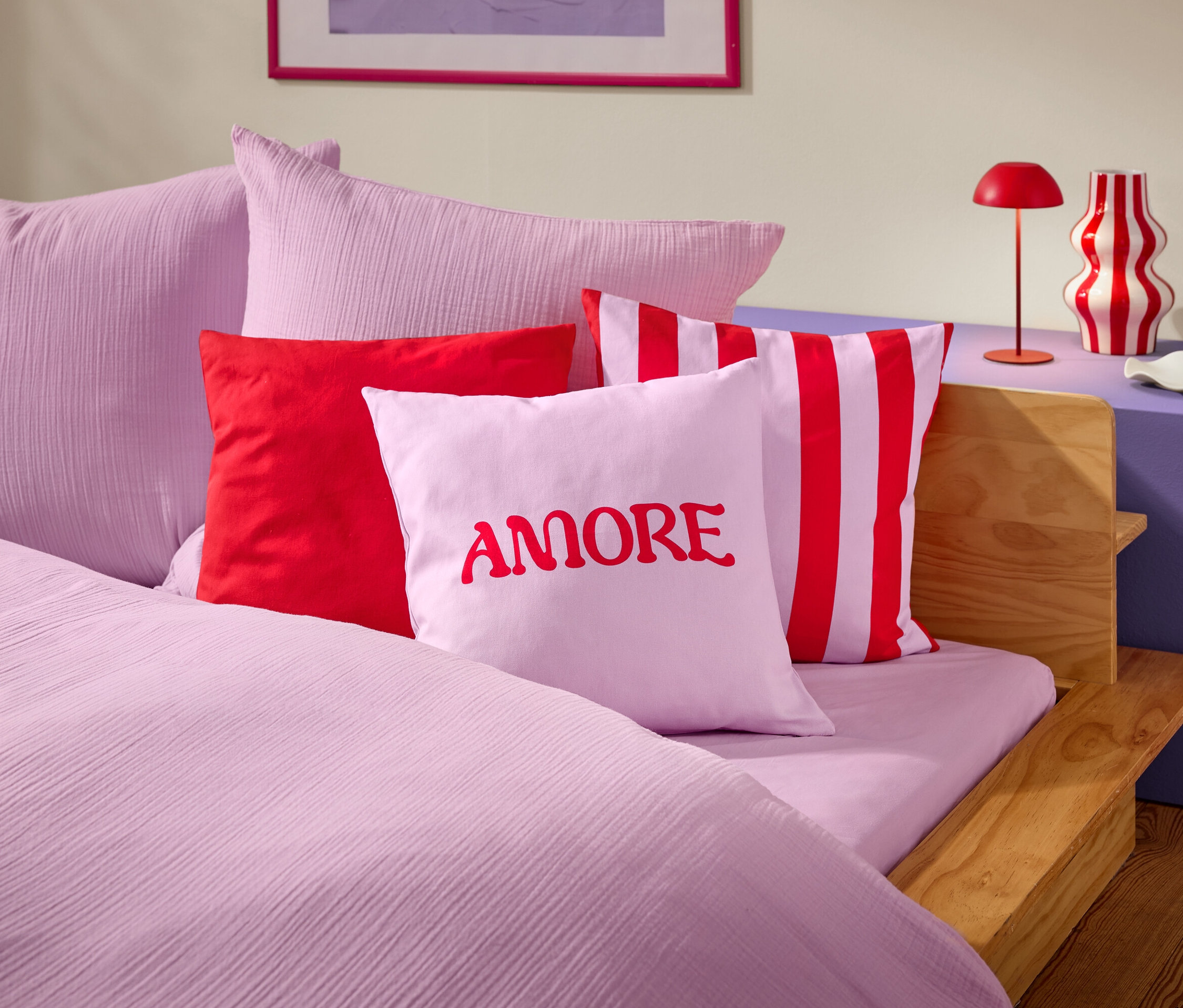 Detailný záber postele s ružovou posteľnou bielizňou a vankúšmi. Na jednom z vankúšov je červeným písmom napísané slovo AMORE.