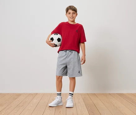 Usmievavý chlapec drží futbal v náručí, má na sebe červené tričko a sivé šortky.