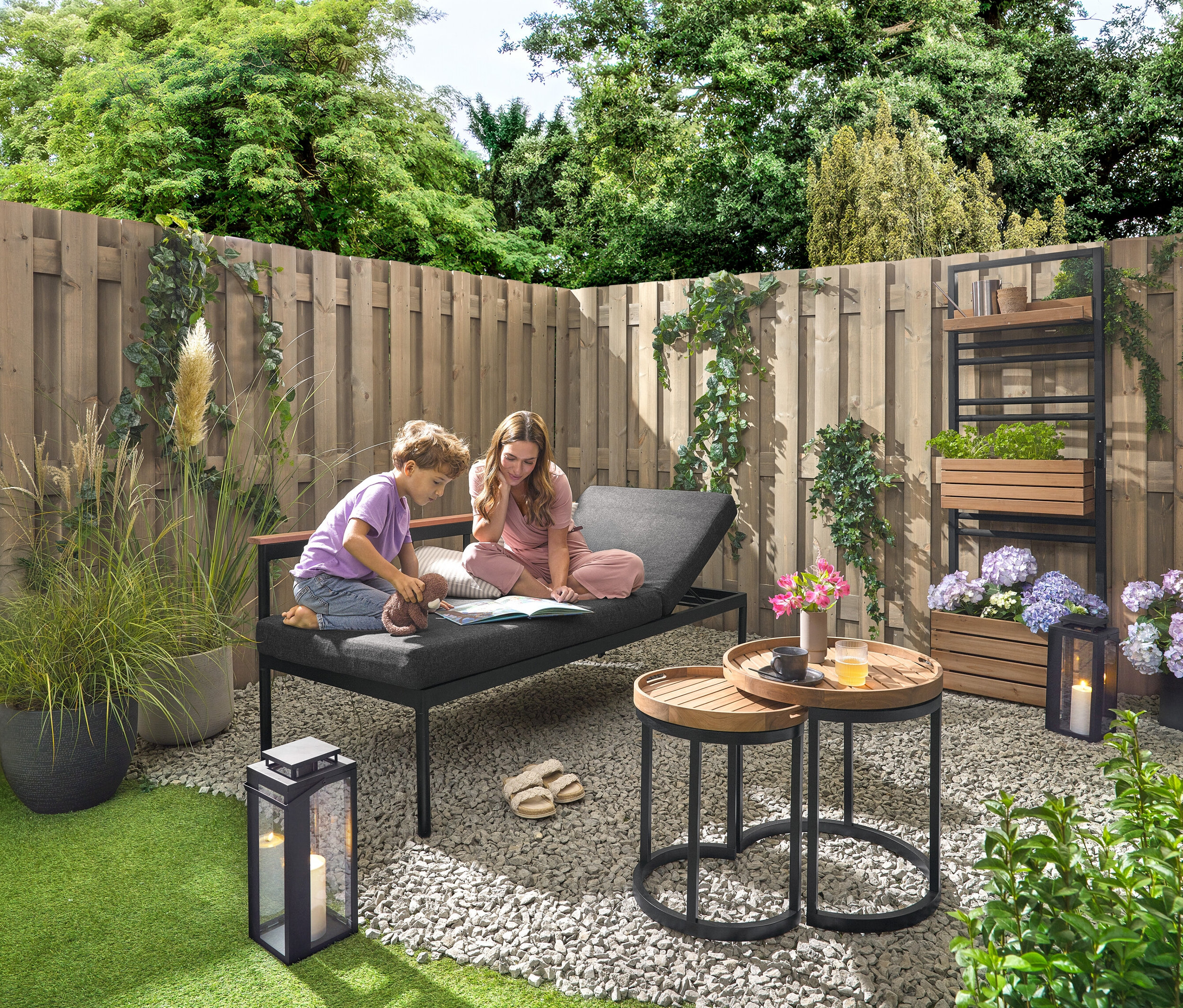 Dieťa a žena sedia na záhradnej lavici 2 v 1 ‒ ležadle »Elin« a čítajú si knihu. V pozadí sa nachádza stena s rastlinami s flexibilne zavesiteľnými policami »Elin«. Vpravo stojí vonkajšia lampáš so sviečkou. K dispozícii sú aj 2 záhradné príručné stolíky s odnímateľnými podnosmi, Ø cca 40 cm a 50 cm, ako aj 2 vonkajšie vankúše s ľanovou štruktúrou.