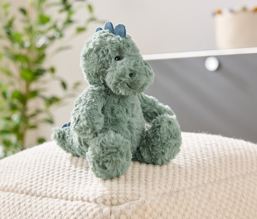 Zelený plyšový dinosaurus sedí na bielej štruktúrovanej stoličke. V pozadí je rastlina a skriňa.