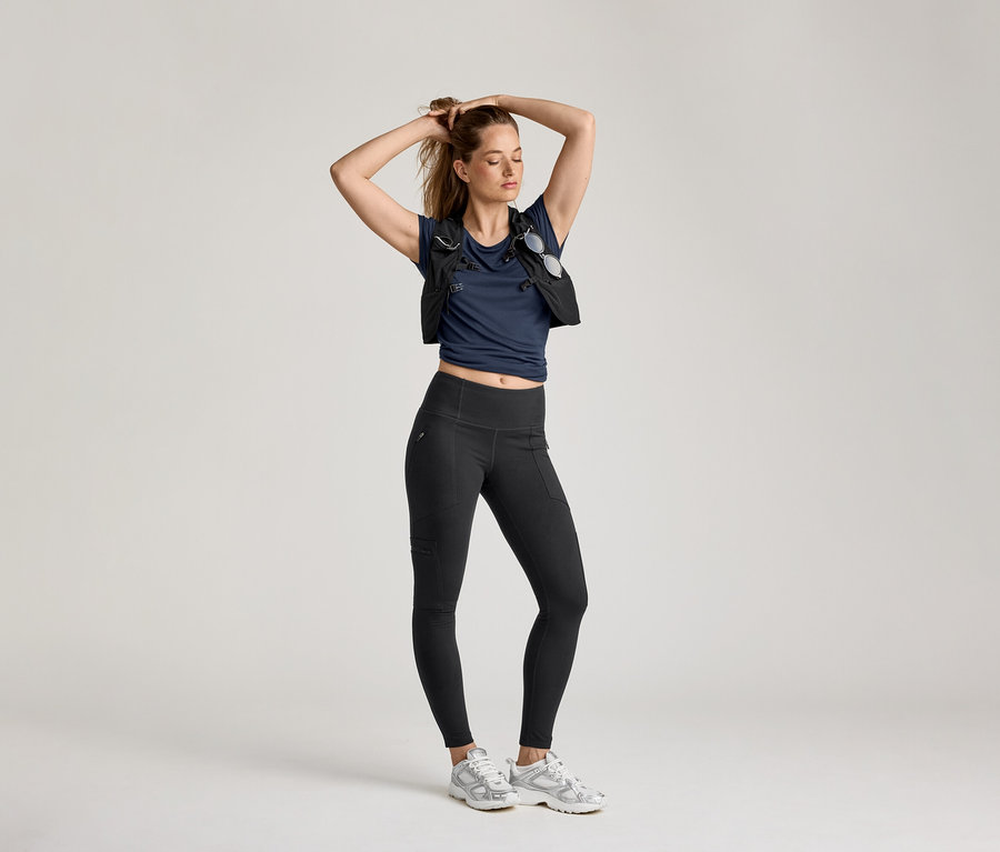 Žena pózuje v outdoorových legínsach, tmavomodrom tričku a veste na svetlom pozadí.