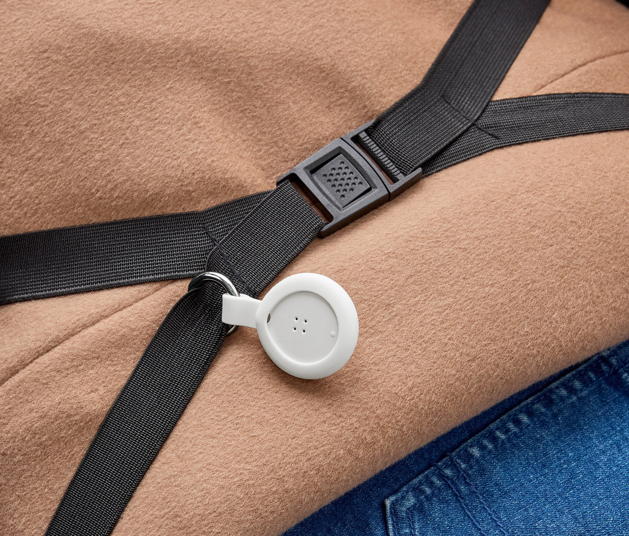 Biele, kruhové zariadenie s tromi bodkami je pripevnené k čiernemu popruhu s kovovým krúžkom. Popruh s čiernou plastovou prackou leží na svetlohnedých látkach a modrom džínsovom materiáli.