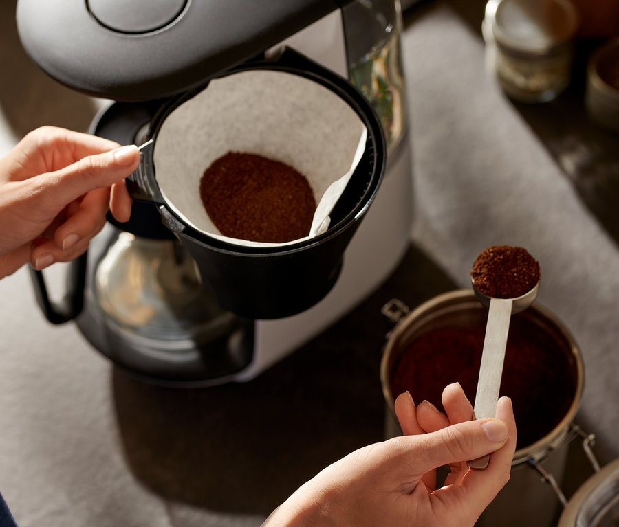 Žena pridáva kávu do bieleho kávovaru na prekvapkávanú kávu »Let's Brew«.