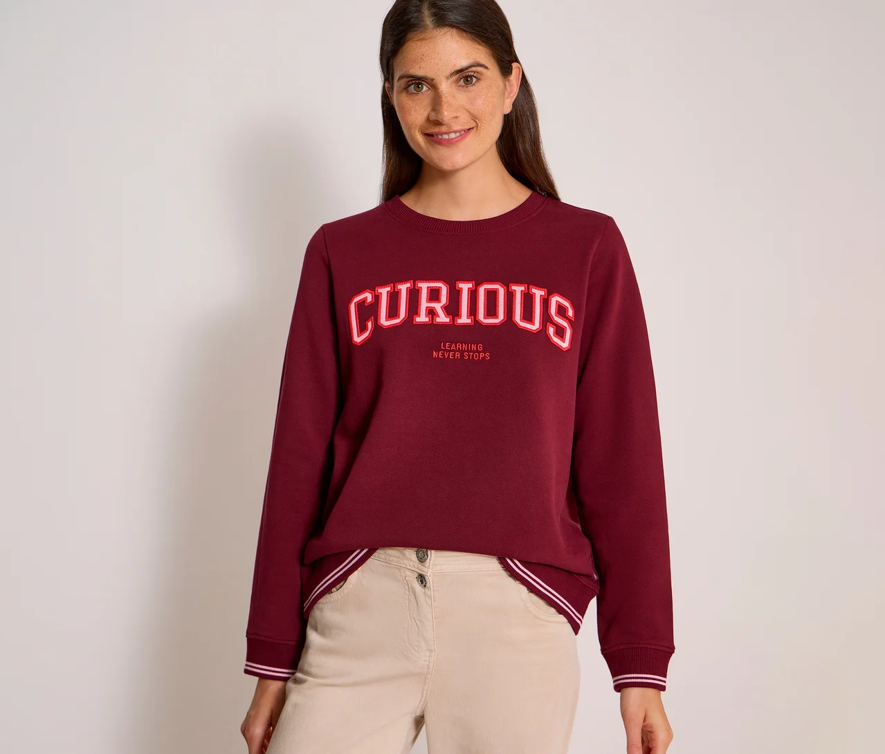Usmievavá žena s tmavými vlasmi má na sebe bordovú mikinu CECIL s nápisom „CURIOUS“ v červeno-bielych písmenách a „LEARNING NEVER STOPS“ pod ním, skombinovanú so svetlobéžovými nohavicami.