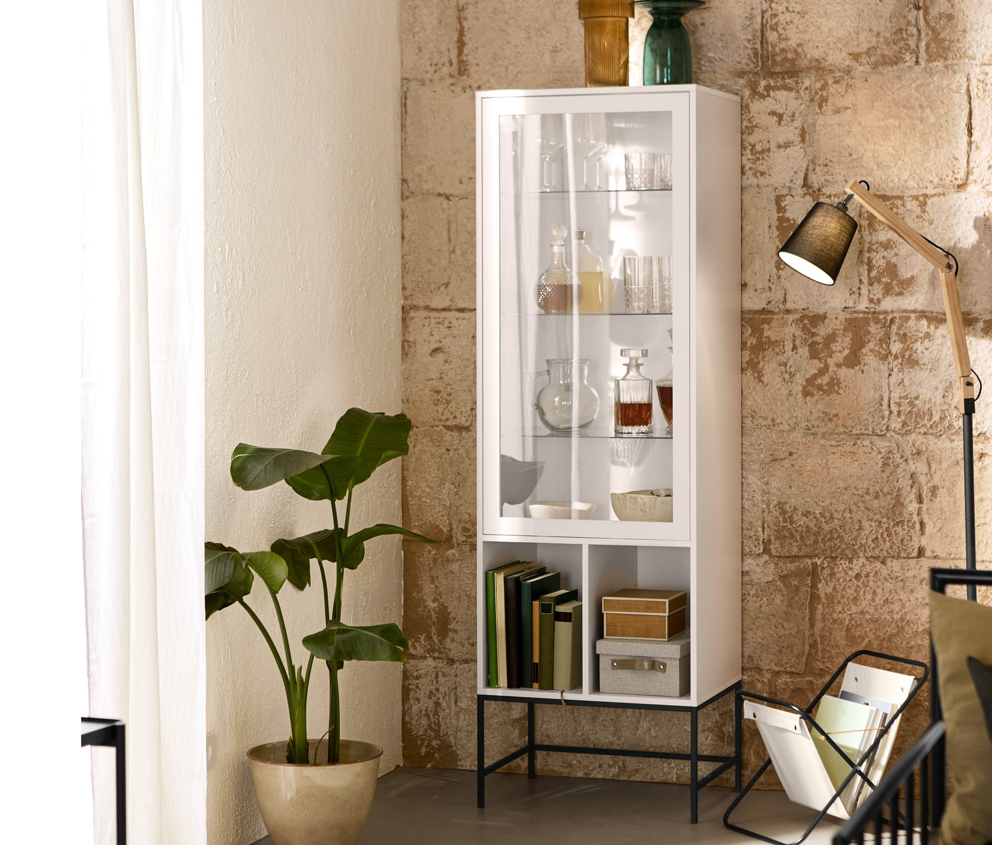 Vitrína so sklenenými policami stojí vedľa stojana na časopisy a stojacej lampy s textilným tienidlom.