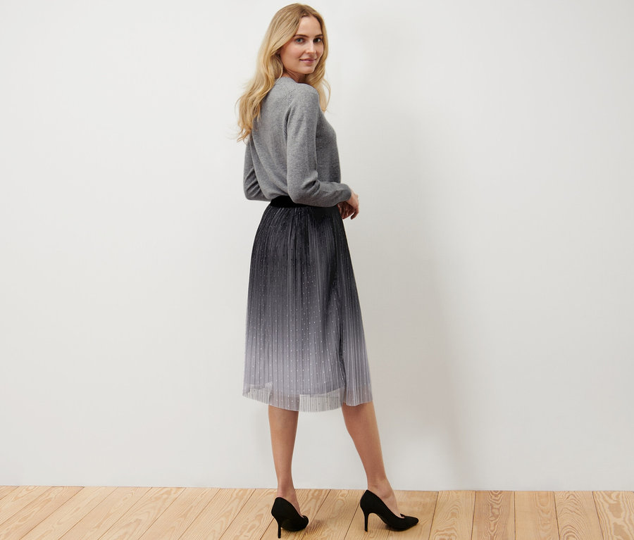 Žena pózuje a má na sebe plisovanú tylovú sukňu s farebným prechodom, sivý sveter a čierne topánky na podpätku.