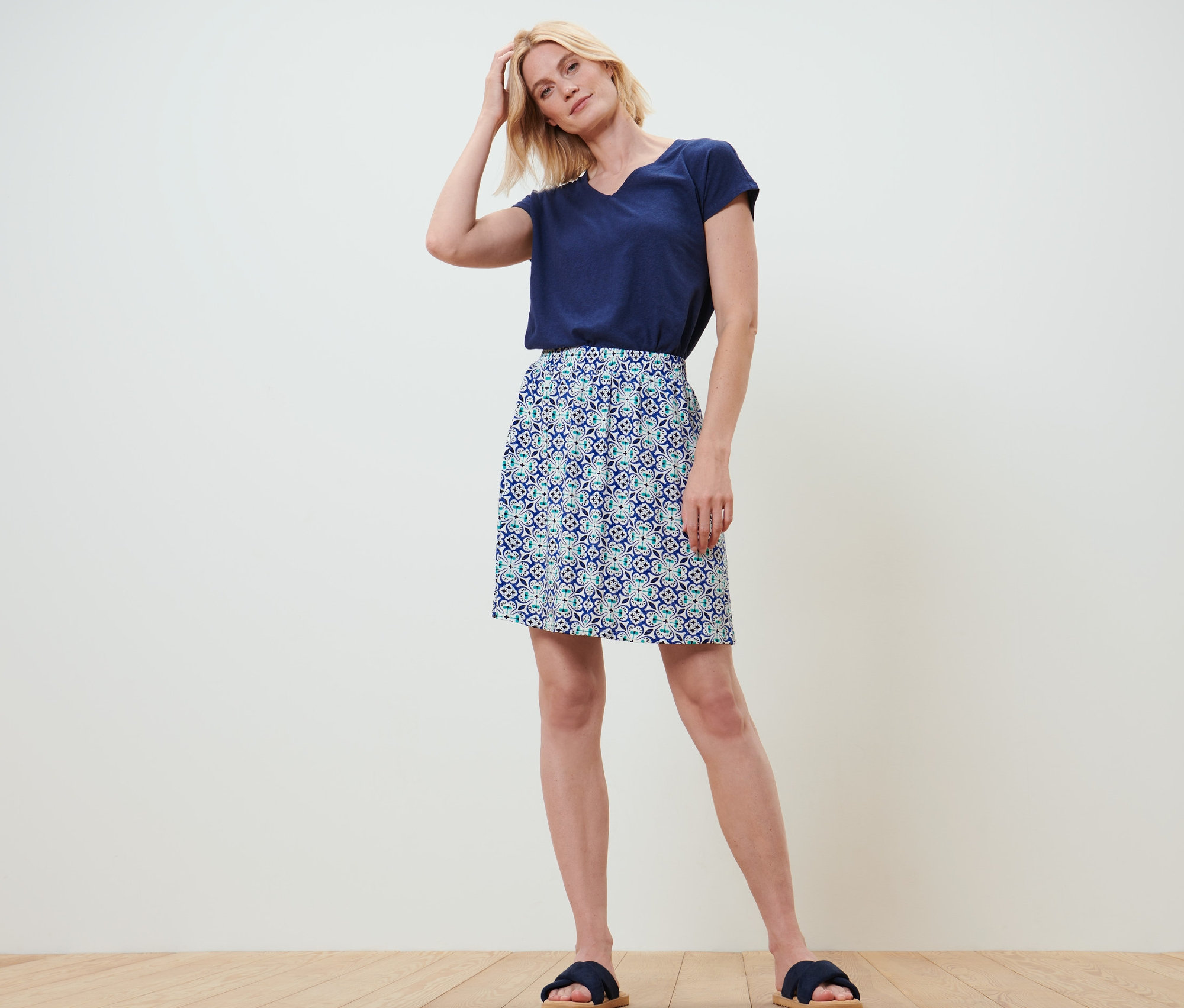 Blondínka stojí a má ruku vo vlasoch. Oblečená je do tmavomodrého trička a džersejovej sukne s celoplošnou potlačou.