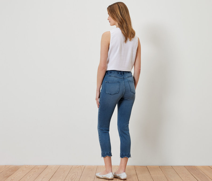 Žena stojaca chrbtom k fotoaparátu má na sebe sedemosminové tregínsy s ažúrovou výšivkou, biely top a strieborné baleríny.