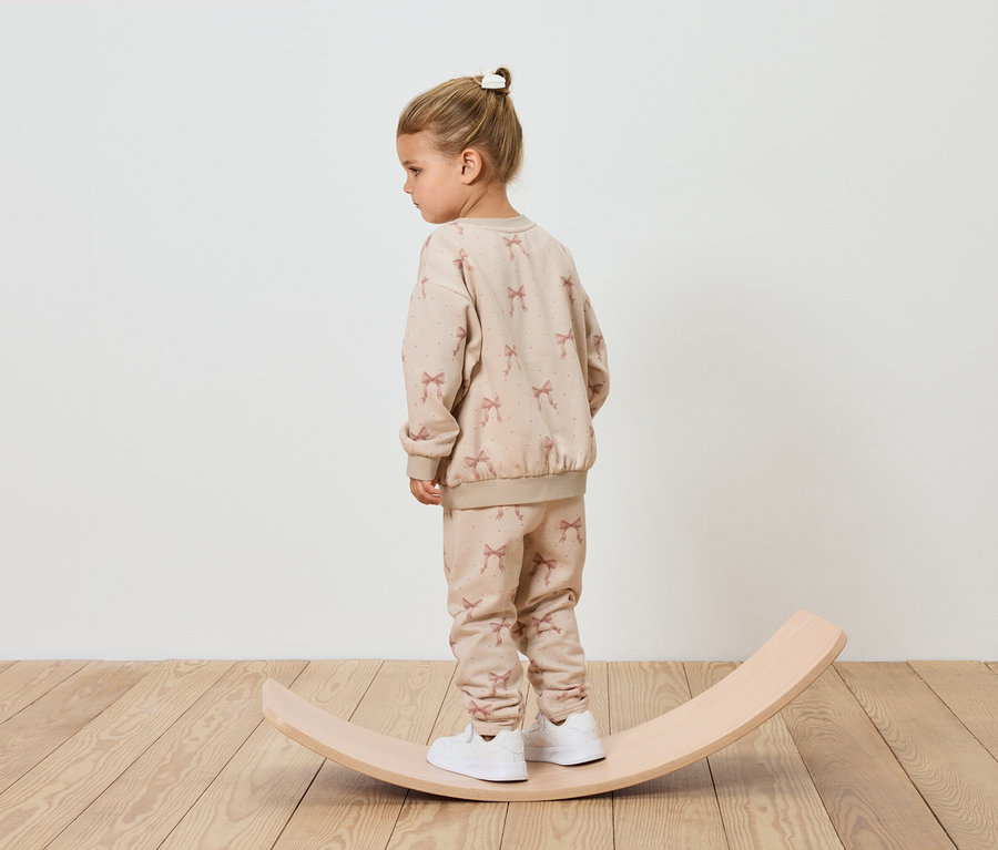 Dievča v béžovej teplákovej súprave stojí na drevenej balančnej doske.