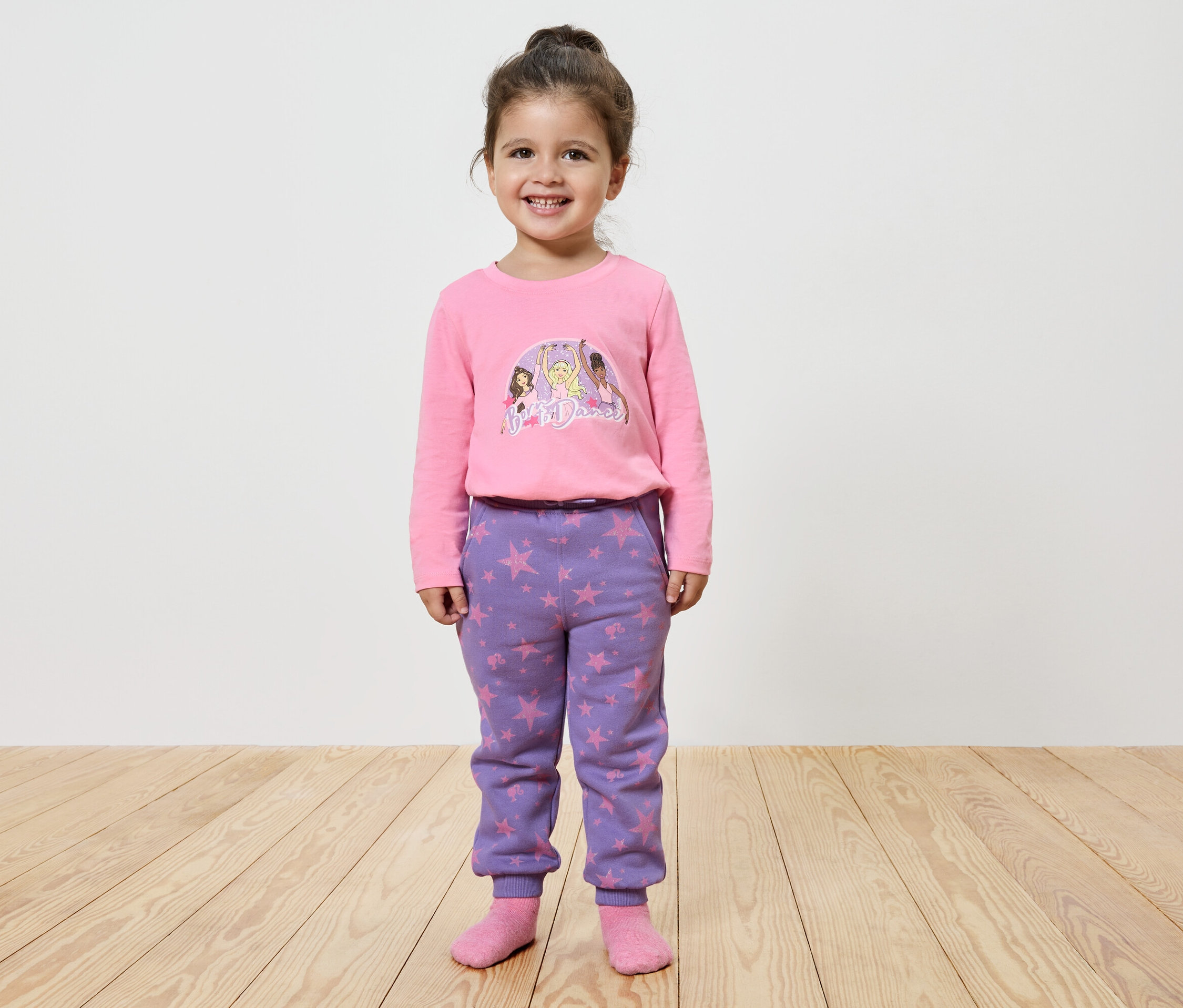 Usmievajúce sa dievčatko pózuje v ružovom tričku s potlačou a fialových nohaviciach s hviezdami.