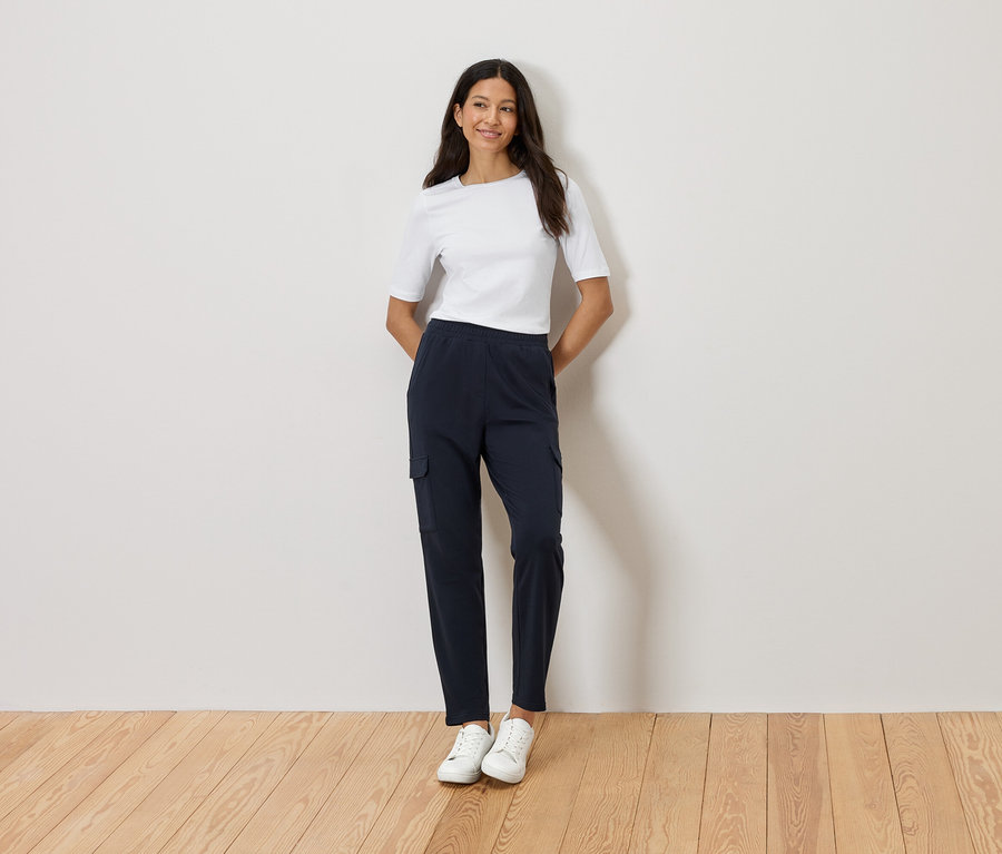 Žena stojí so založenými rukami pred stenou, má na sebe biele tričko, tmavomodré cargo nohavice a biele tenisky.