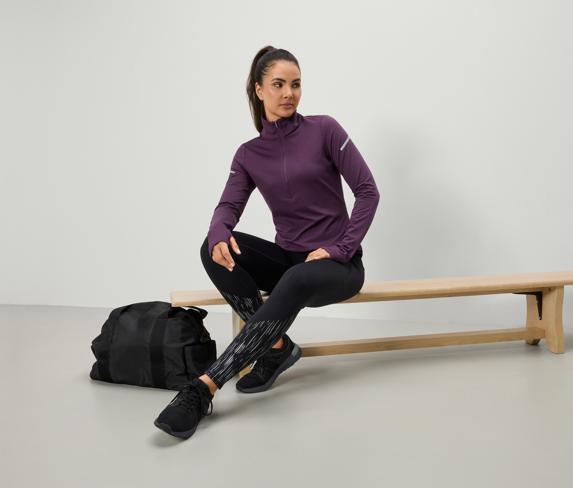 Žena sedí na lavičke v purpurovej športovej košeli a čiernych legínach, vedľa nej leží čierna športová taška.