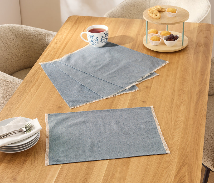 Dve modré prestierania s bielym okrajom na drevenom stole, vedľa nich šálka, taniere a etažér s jedlom.