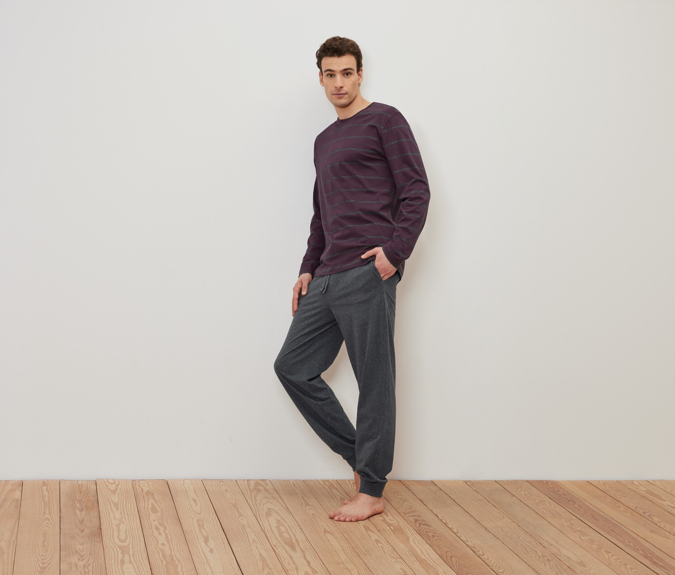 Muž stojí bosý na drevenej podlahe a má na sebe fialové pruhované tričko s dlhým rukávom a sivé tepláky.