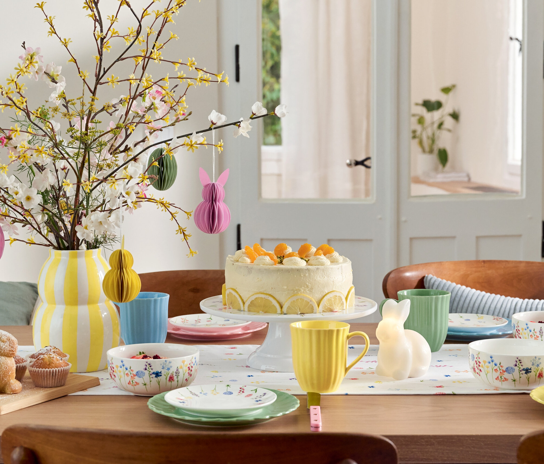 Jedálenský stôl s veľkonočnou výzdobou, koláčom, riadom a vázou s kvetmi.