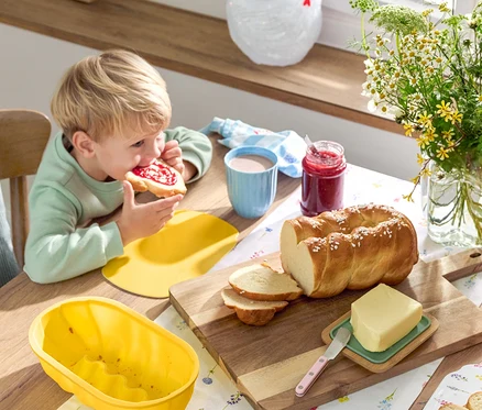 Blond chlapec je džemový chlieb pri stole s pletenkou, maslom, džemom, riadom a kvetmi.