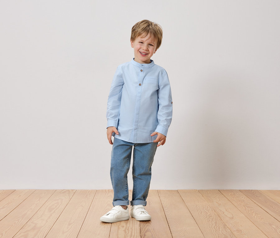 Usmievavý chlapec s blond vlasmi stojí pred bielou stenou. Má na sebe svetlomodrú košeľu, džínsy a biele topánky.