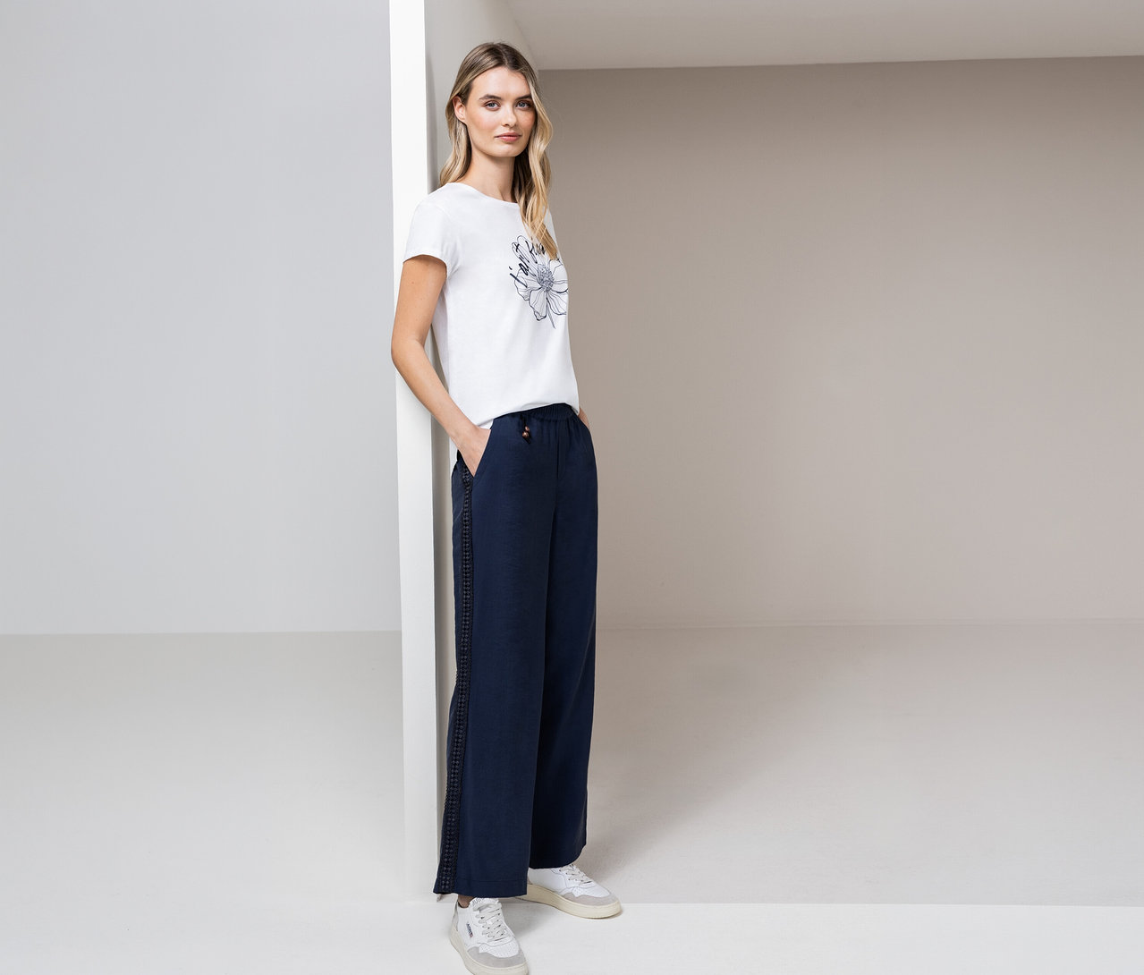 Žena pózuje v tričku Street One, modrých nohaviciach a bielych teniskách v svetlej miestnosti.