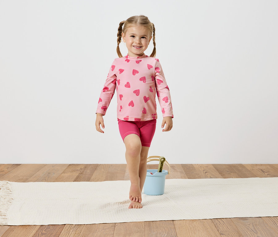 Dievča s vrkočmi, má na sebe ružový top a šortky so srdiečkami, balansuje na bielom koberci vedľa svetlomodrého vedra.