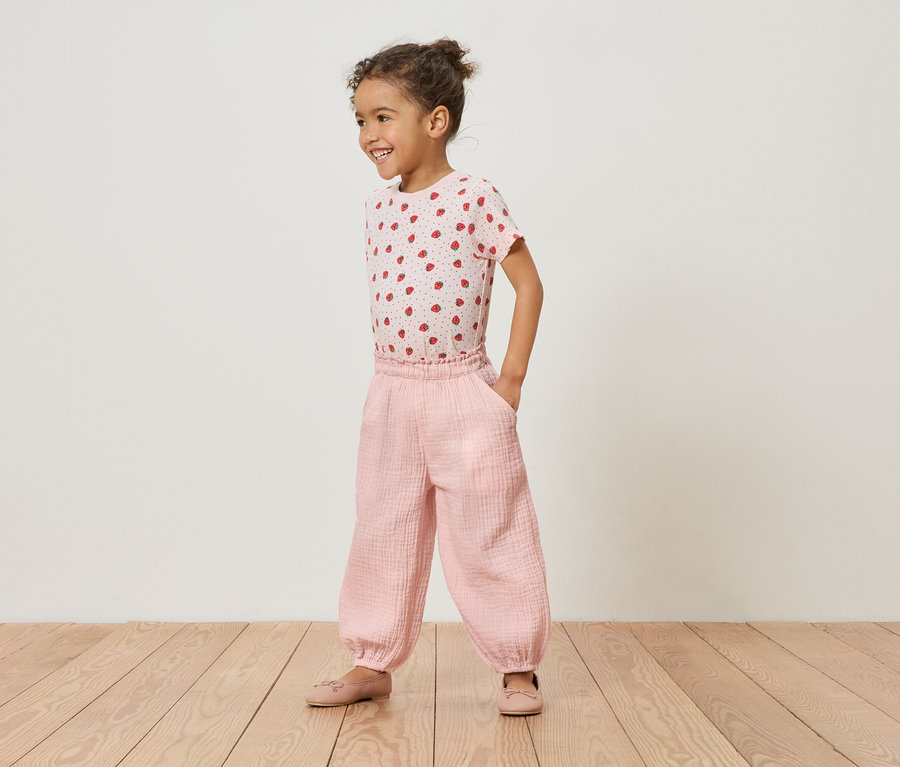 Dievča stojí na drevenej podlahe v ružovom tričku s jahodami a ružových nohaviciach.