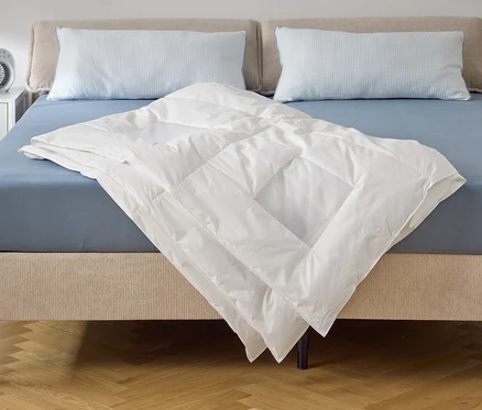 Na posteli s modrou plachtou a dvoma vankúšmi leží biela prikrývka. Na nočnom stolíku stojí ventilátor.