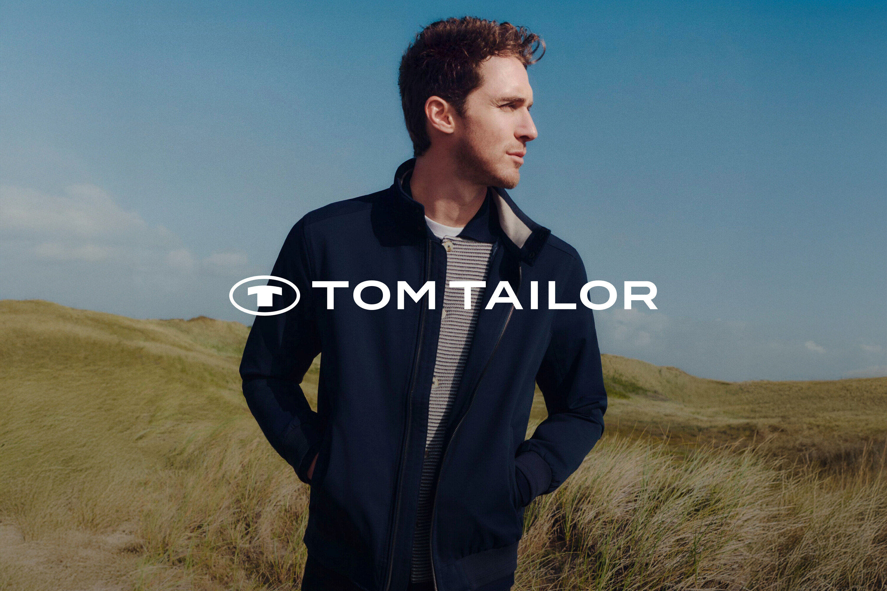 Muž v tmavomodrej bunde a pruhovanom tričku pred trávnatou krajinou a modrou oblohou.