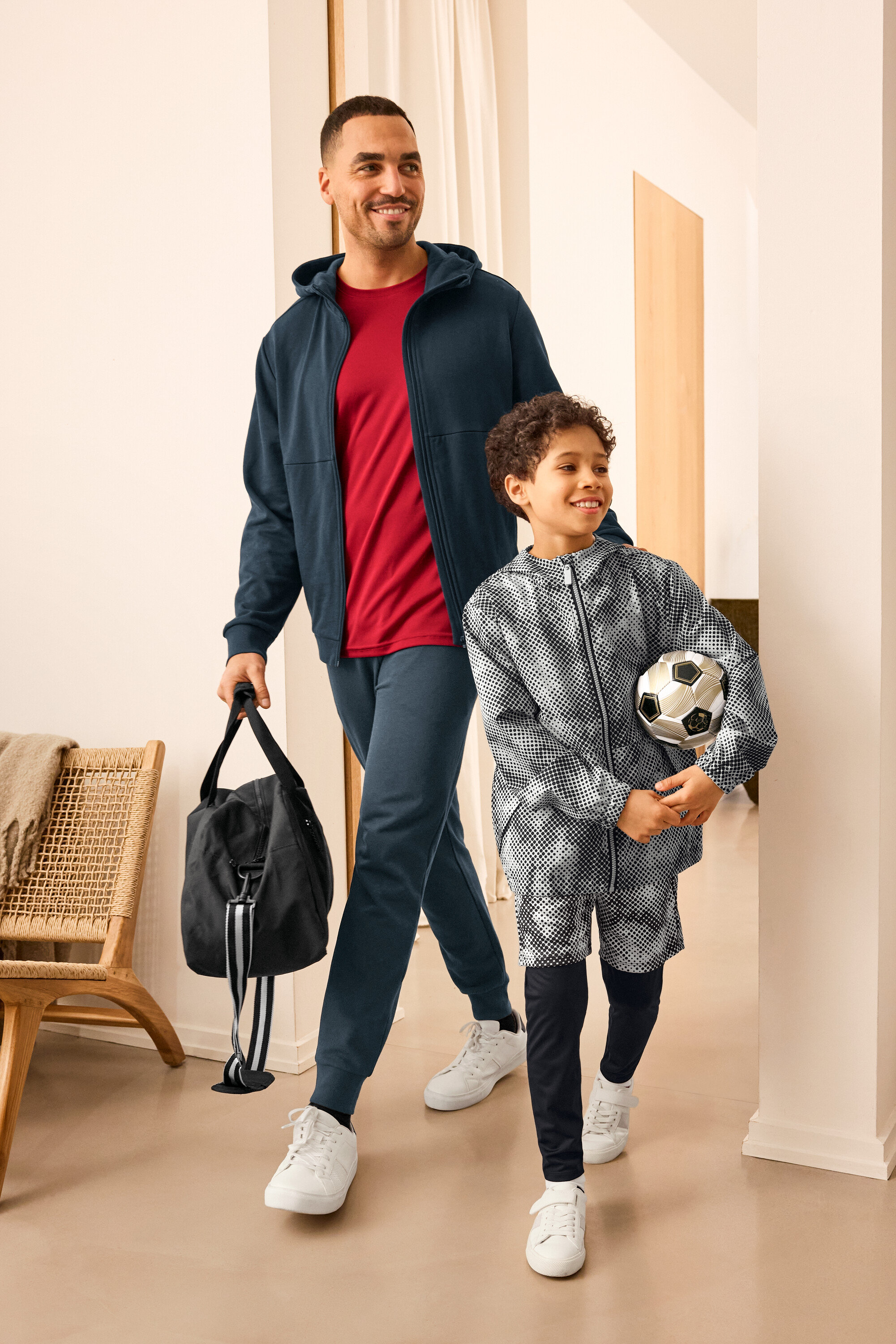 Muž so športovou taškou a dieťa s futbalom, obaja majú na sebe športové oblečenie. Muž má na sebe Sweatjacke a Sweathose. Dieťa má na sebe Kinder-Windbreaker-Jacke.