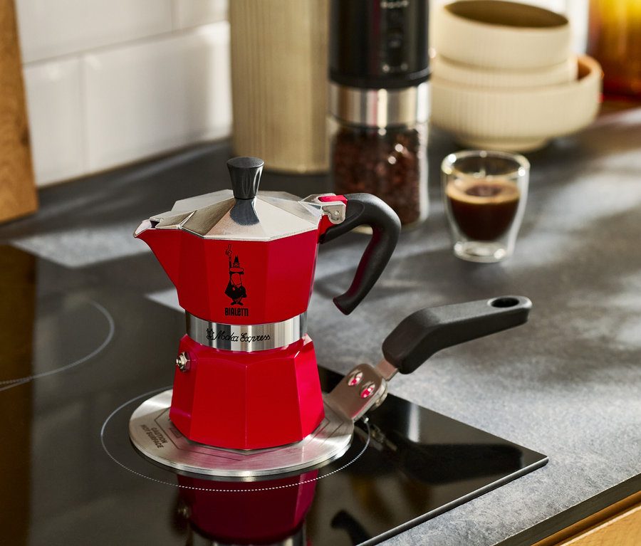 Červený kávovar na espresso stojí na varnej doske vedľa šálky espressa.
