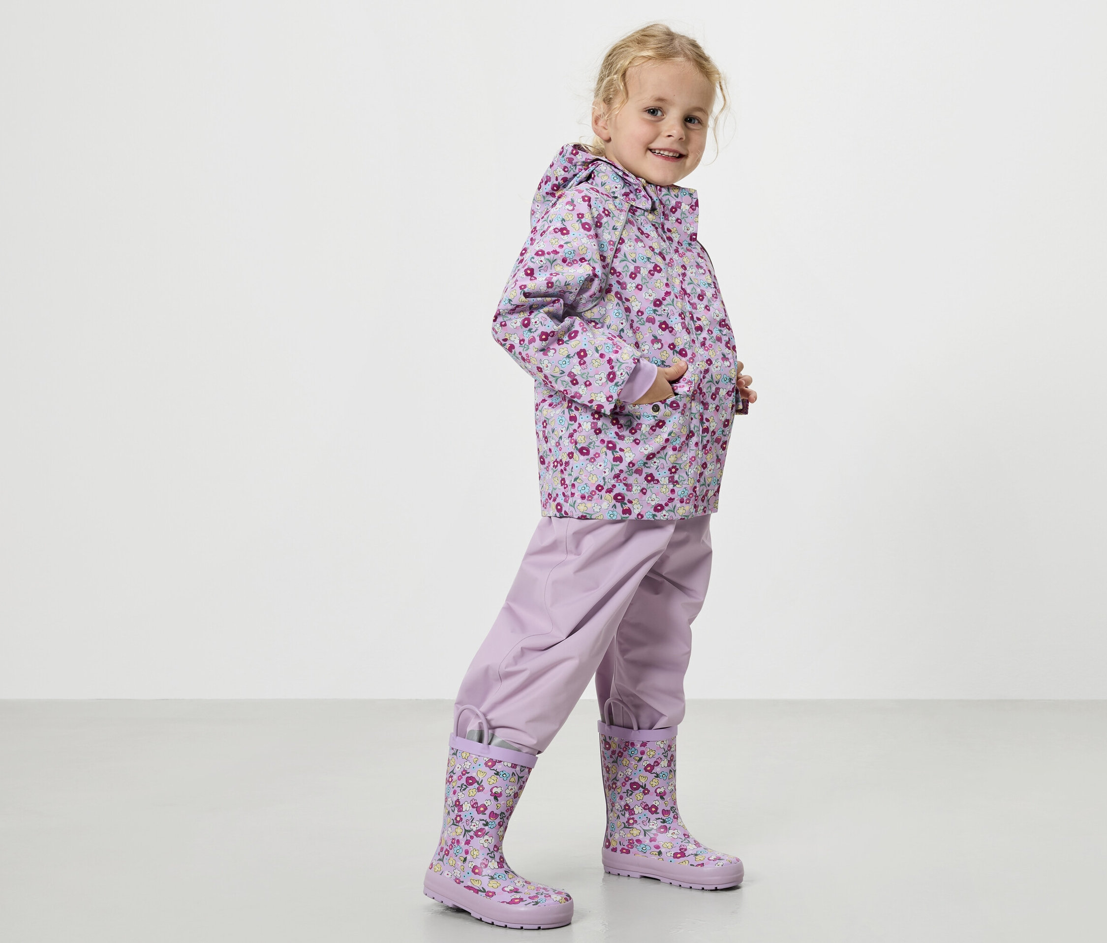 Blondínka má na sebe fialový pršiplášť s kvetinovým vzorom, fialové nohavice do dažďa a zodpovedajúce gumáky.