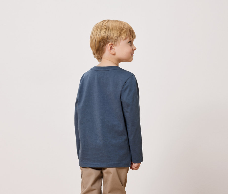 Blond chlapec stojí chrbtom k fotoaparátu a má na sebe modré tričko s dlhým rukávom a khaki nohavice.