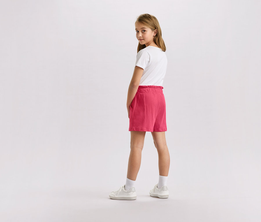 Dievča s blond vlasmi v bielom tričku, ružových šortkách a bielych teniskách, pozerajúce sa do kamery.