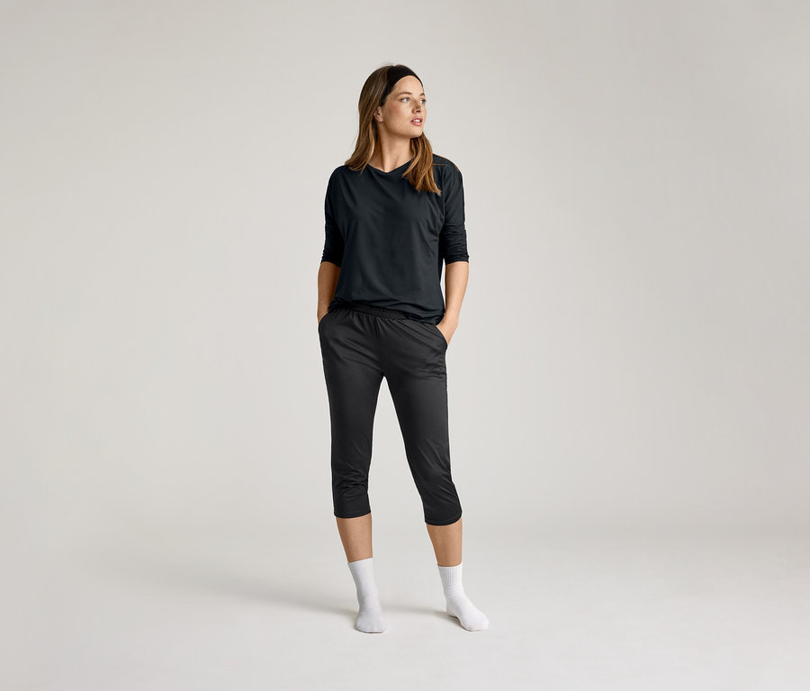 Žena v čiernom 3/4 športovom tričku a tmavých nohaviciach, ruky vo vreckách, biele ponožky, na svetlom pozadí.