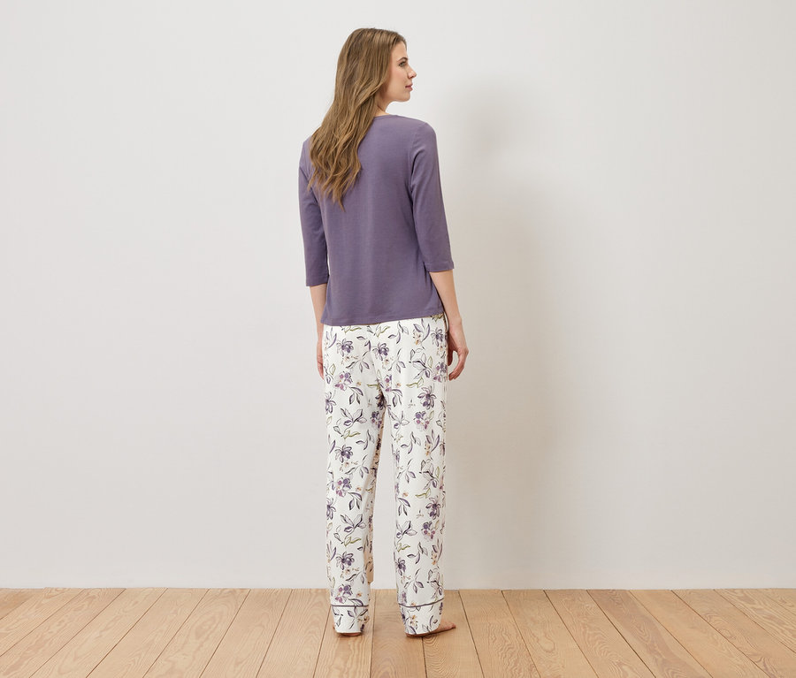 Žena stojí chrbtom k fotoaparátu a má na sebe fialový pyžamový top a biele pyžamové nohavice s kvetinovým vzorom.