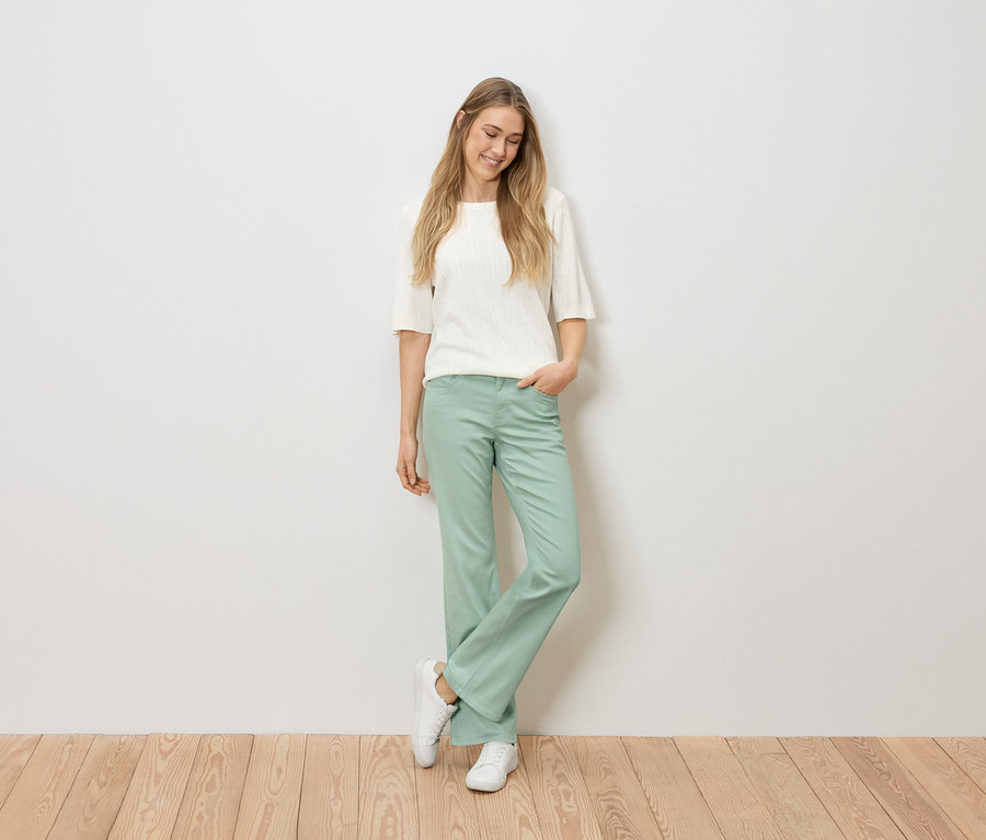 Žena s blond vlasmi v bielom tope, zelených nohaviciach a bielych teniskách stojí na drevenej podlahe pred bielou stenou.