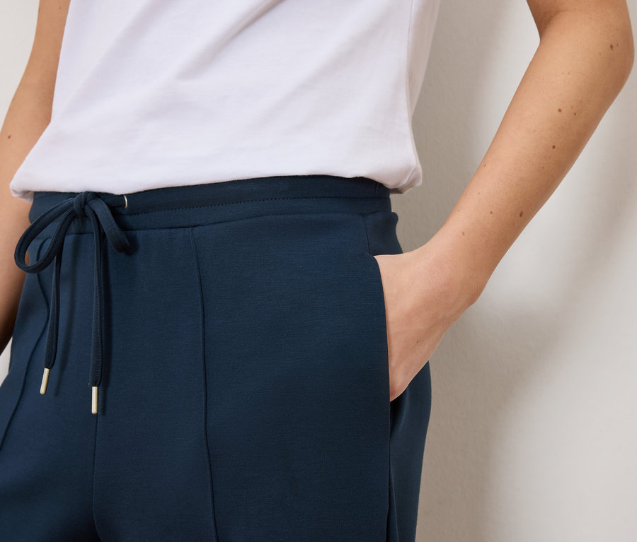 Zblízka žena v tmavomodrých teplákoch so šnúrkou a zlatými detailmi a bielom tričku.