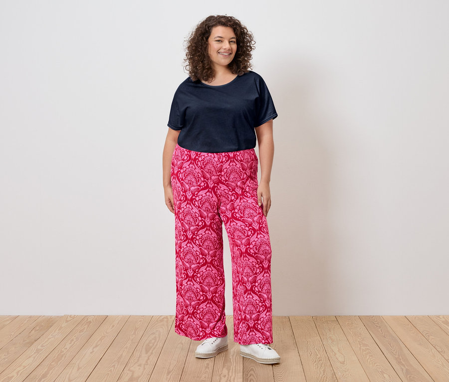 Žena s kučeravými vlasmi má na sebe tmavomodré tričko, ružové vzorované nohavice a biele tenisky.