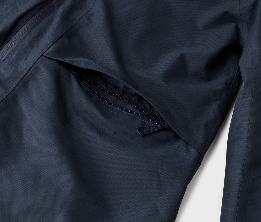 Detail tmavomodrej bundy, zobrazujúci čiastočne otvorenú vrecku na zips vpredu a vertikálny zips na ľavej strane.