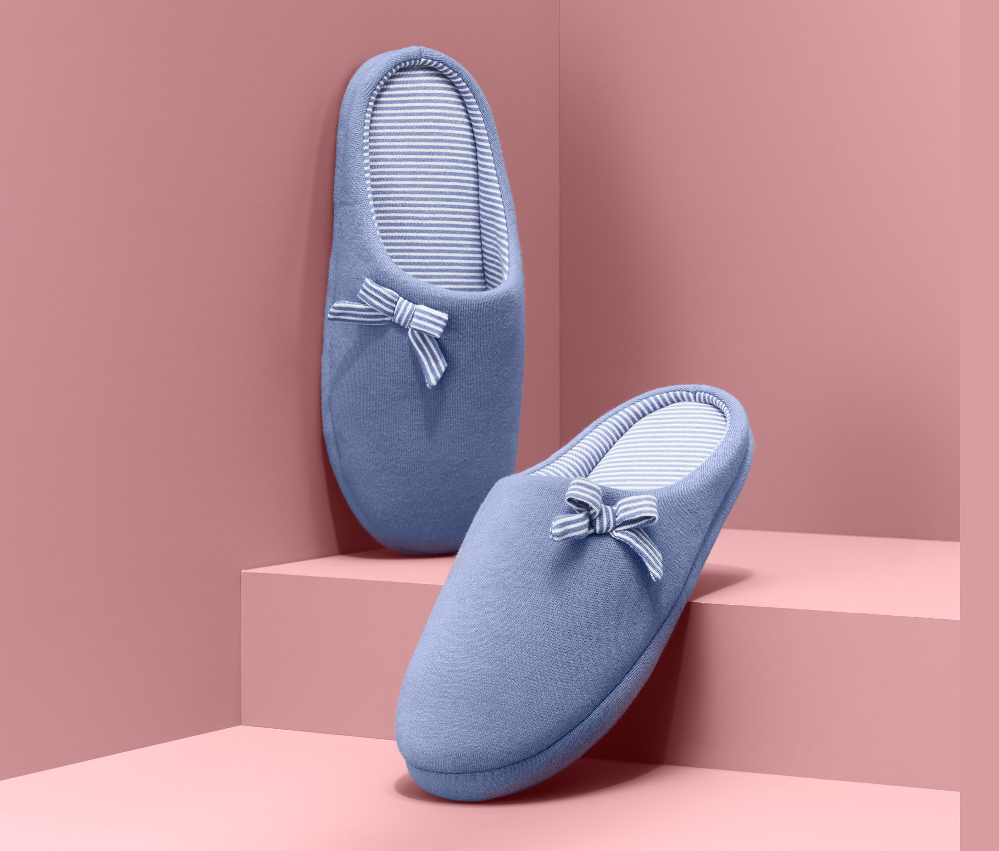 Dve modré papuče s prúžkovaným vnútrom a mašľou stoja na ružových schodoch.